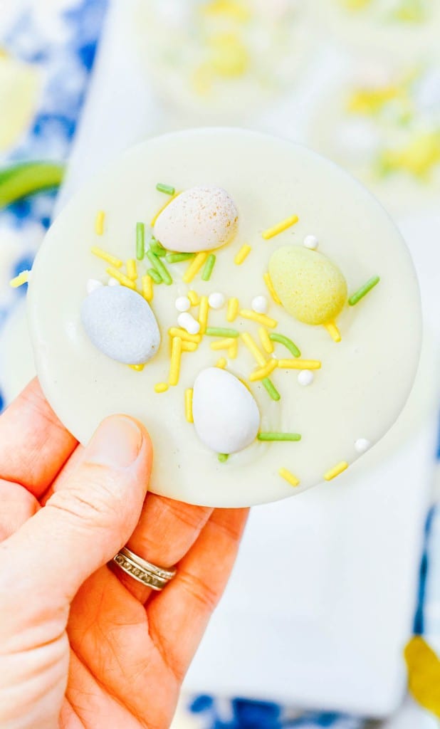 White chocolate disks topped with small pastel eggs and sprinkles sit on a tray. Nearby, there are yellow tulips and a wooden bunny figure on a blue floral cloth.