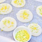 Round white cheese slices on a sheet, each topped with yellow and green sprinkles. A small bowl filled with the same sprinkles is placed nearby.