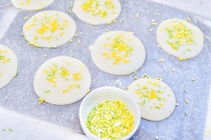 Round white cheese slices on a sheet, each topped with yellow and green sprinkles. A small bowl filled with the same sprinkles is placed nearby.
