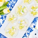 White chocolate circles topped with colored candy eggs and sprinkles are arranged on a rectangular plate. Yellow tulips and a decorative rabbit napkin are nearby on a blue patterned cloth.