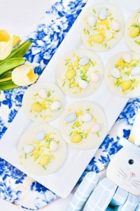White chocolate circles topped with colored candy eggs and sprinkles are arranged on a rectangular plate. Yellow tulips and a decorative rabbit napkin are nearby on a blue patterned cloth.
