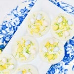 White chocolate discs topped with pastel-colored candy eggs and yellow sprinkles on a white plate, placed on a blue patterned cloth near yellow tulips.