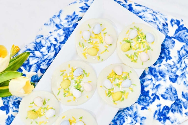 White chocolate discs topped with pastel-colored candy eggs and yellow sprinkles on a white plate, placed on a blue patterned cloth near yellow tulips.