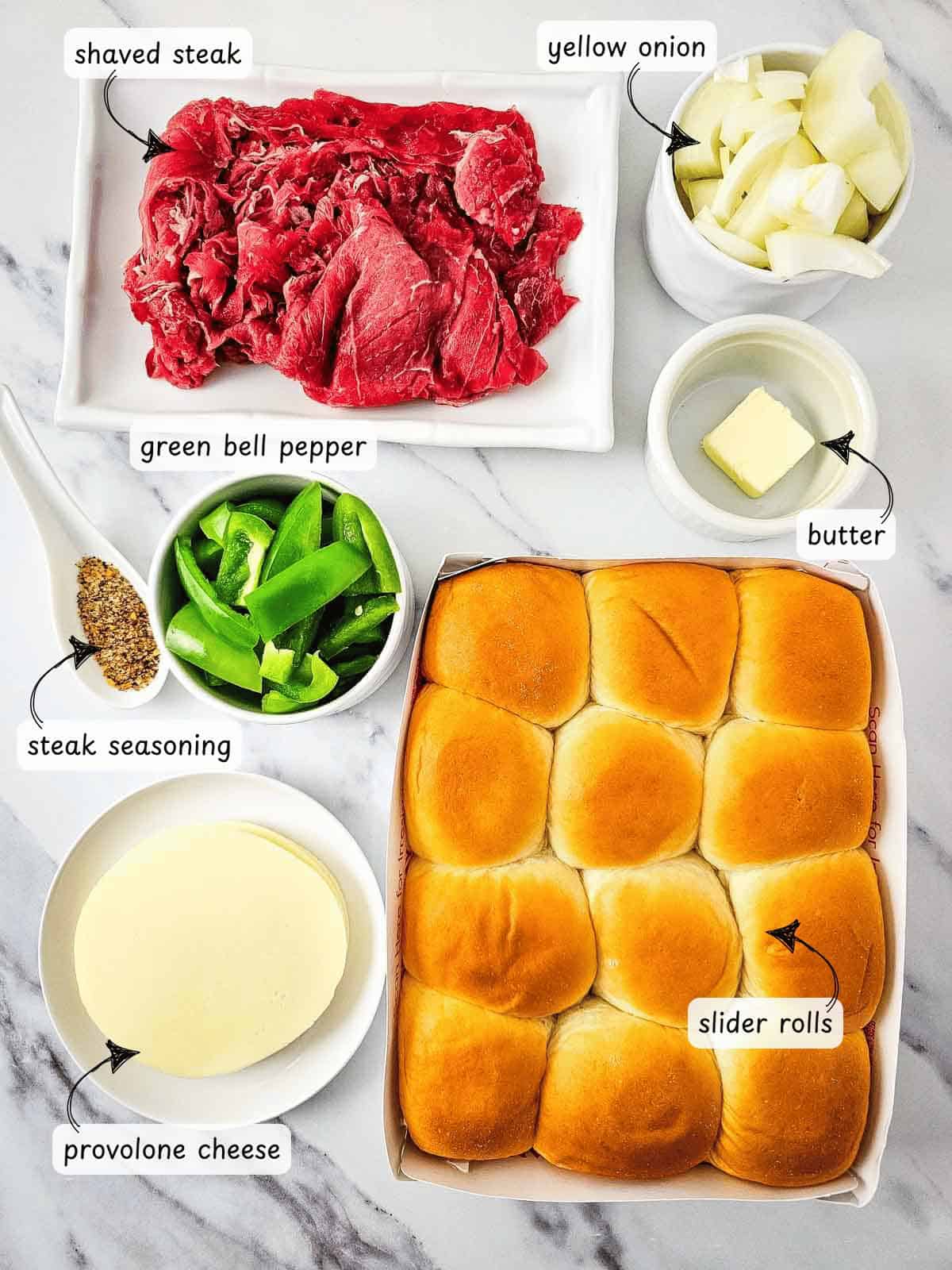 Ingredients for sliders: shaved steak, yellow onion, green bell pepper, steak seasoning, provolone cheese, butter, and slider rolls on a marble surface.
