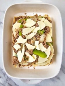 Unbaked casserole with layers of sliced onions, green peppers, shredded meat, and cheese in a white baking dish.
