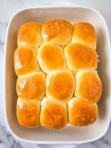 A baking dish with twelve golden brown dinner rolls, brushed with butter, on a marble surface.