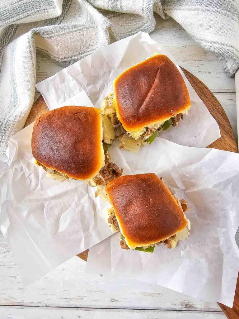 Three pulled pork sandwiches on a wooden platter, placed on crumpled parchment paper, with a striped cloth in the background.