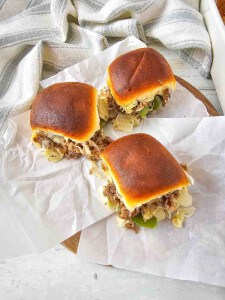 Three beef sliders with cheese, onions, and peppers on a white paper-lined surface.