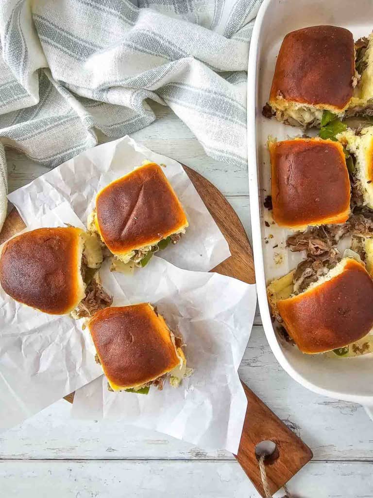 Four sliders on parchment paper next to a dish with more sliders, on a wooden board. Rolls are golden brown and filled with shredded meat and cheese. Striped cloth in the background.