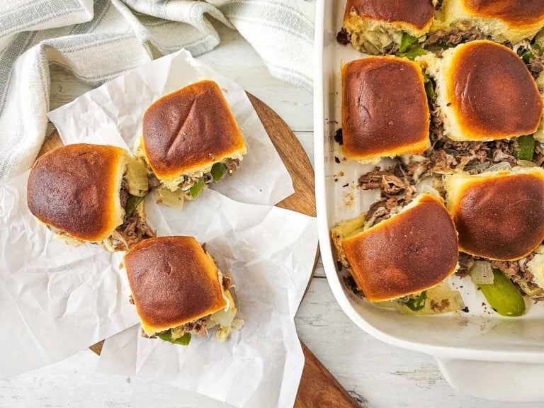 Sliders with meat, cheese, and green peppers are on parchment paper and in a baking dish. A striped cloth is in the background.