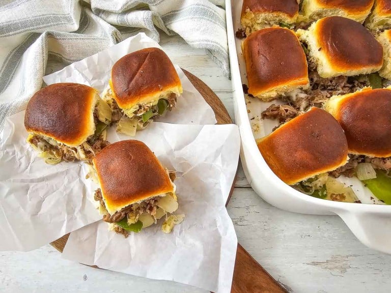 Sliders with meat, onions, and green peppers on white parchment paper, next to a baking dish with more sliders. A striped cloth is partially visible in the background.