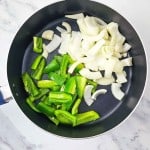 A frying pan on a marble surface contains sliced green bell peppers and onions.