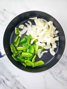 A frying pan on a marble surface contains sliced green bell peppers and onions.