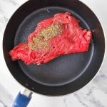 Raw beef steak seasoned with pepper and spices in a nonstick frying pan on a marble surface.