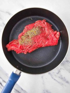 Raw beef steak seasoned with pepper and spices in a nonstick frying pan on a marble surface.