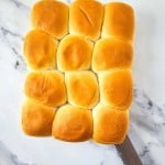 A dozen freshly baked dinner rolls arranged in a 3x4 grid on a marble surface, with a serrated knife placed at the bottom right corner.