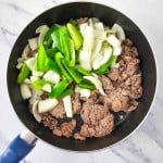 Ground beef, sliced green bell peppers, and chopped onions in a black skillet on a marble surface.