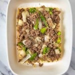 A white dish with a layer of shredded meat, sliced green bell peppers, and onion pieces on a marble countertop.