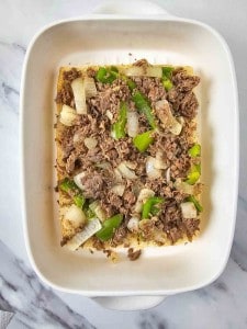 A white dish with a layer of shredded meat, sliced green bell peppers, and onion pieces on a marble countertop.