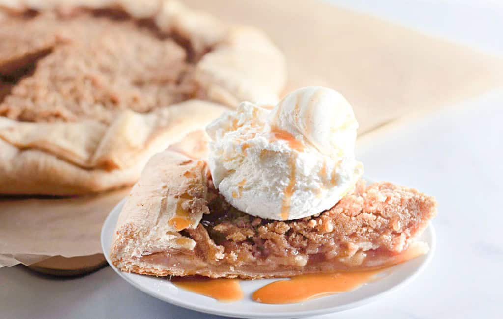 A slice of apple pie from our favorite apple recipes collection, topped with a scoop of vanilla ice cream and drizzled with caramel sauce on a white plate, while the whole pie waits enticingly in the background.