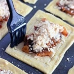 A pastry brush glazes raw puff pastry for a delectable apple crumb Danish, topped with a mixture of chopped apples and crumbly streusel on a baking tray.