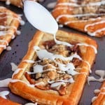 A spoon drizzles white icing over rectangular apple crumb danishes, each topped with golden crumbles on a dark baking sheet.