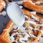A spoon drizzles white icing over a rectangular apple crumb Danish with a crumbly topping, resting on a baking sheet.