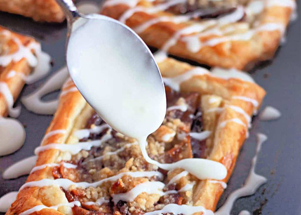 A spoon drizzling white icing over a rectangular apple crumb Danish with a crumbly topping on a tray.