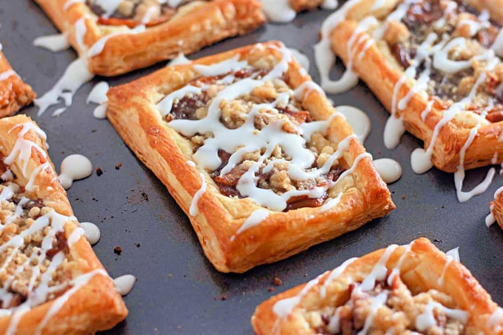 Rectangular apple crumb danishes with a flaky crust are topped with a crumbly filling and a white icing drizzle, artfully arranged on a dark baking sheet.