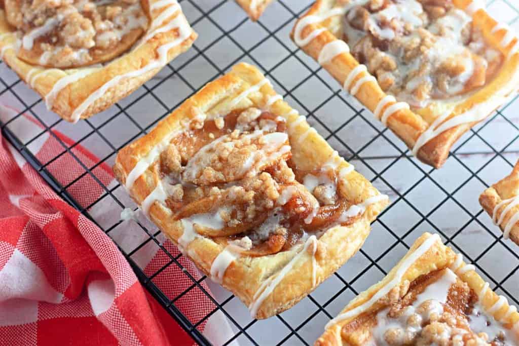 Rectangular apple crumb danishes with streusel topping and white icing are cooling on a wire rack. A red and white checkered cloth is partially visible.