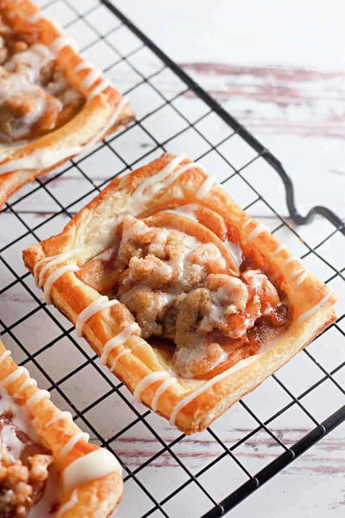 A rectangular apple crumb Danish topped with apple slices, crumb topping, and drizzled icing sits on a black cooling rack.