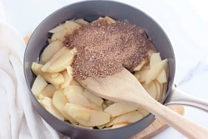 Sliced apples in a pan with brown sugar and spices, reminiscent of an apple crumb Danish, being stirred with a wooden spatula.