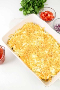 A rectangular baking dish filled with shredded cheese sits on a white surface, surrounded by bowls of chopped tomato, red onion, cilantro, and a measuring cup of red sauce.
