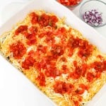 A white baking dish filled with shredded cheese and salsa, next to bowls of chopped tomatoes, red onions, and cilantro on a white surface.