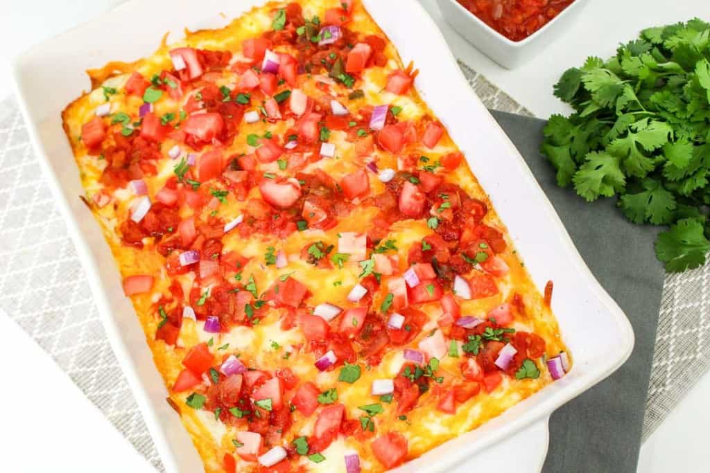 A baked casserole topped with melted cheese, diced tomatoes, red onions, and chopped cilantro, placed in a white baking dish next to fresh cilantro.