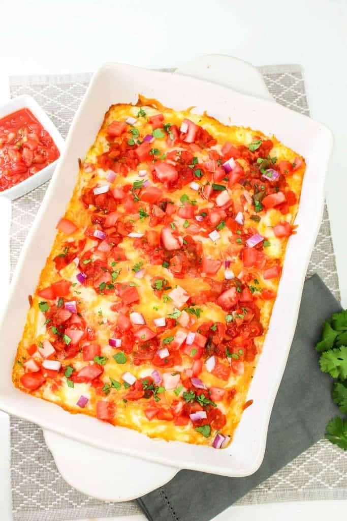 A baked casserole topped with diced tomatoes, red onions, and chopped herbs on a white dish, placed on a gray patterned mat with a small bowl of salsa and fresh cilantro nearby.