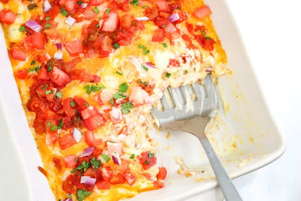 A casserole dish with partially served cheesy enchiladas, topped with diced tomatoes, red onions, cilantro, and salsa. A serving spatula rests inside the dish.