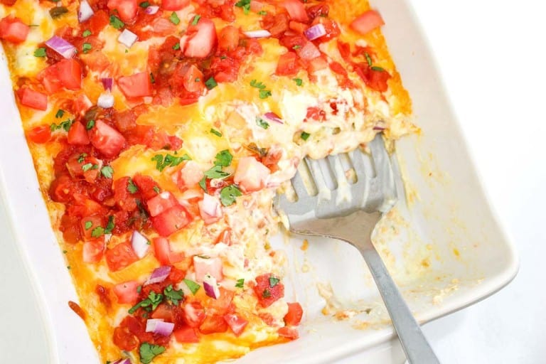 A casserole dish with partially served cheesy enchiladas, topped with diced tomatoes, red onions, cilantro, and salsa. A serving spatula rests inside the dish.