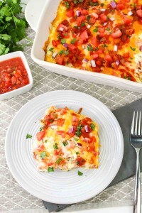 A serving of cheesy enchilada casserole topped with chopped tomatoes, red onions, and herbs on a white plate next to a baking dish and a small bowl of salsa.