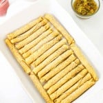 A white baking dish filled with neatly arranged rolled tortillas, surrounded by small bowls of various toppings on a white surface.
