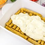 A baking dish filled with rolled tortillas is being topped with a creamy white sauce. Bowls of shredded cheese, diced tomatoes, and chopped red onions are nearby.