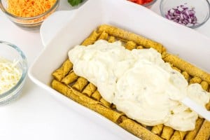 A baking dish filled with rolled tortillas is being topped with a creamy white sauce. Bowls of shredded cheese, diced tomatoes, and chopped red onions are nearby.