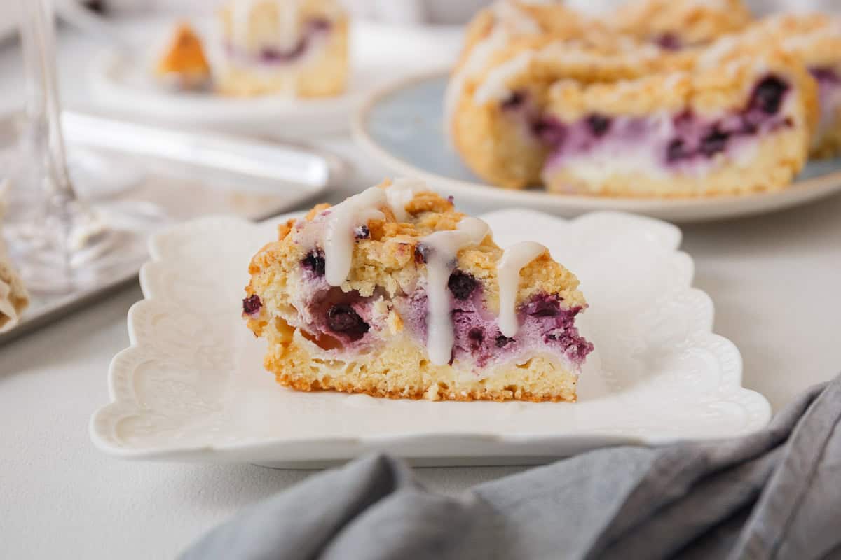 A slice of blueberry coffee cake with white icing rests elegantly on a decorative white plate, displayed on a table where more cake awaits, reminiscent of the indulgence found in cheesecake recipes.