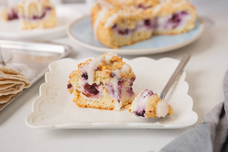 A piece of berry scone with icing rests on a decorative plate, alongside a fork. Another scone, reminiscent of delightful cheesecake recipes, is visible in the background on a blue plate.