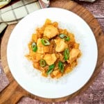A white plate of Crock Pot Hawaiian Chicken curry with diced vegetables and green onions is served over white rice on a wooden board.