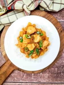 A white plate of Crock Pot Hawaiian Chicken curry with diced vegetables and green onions is served over white rice on a wooden board.