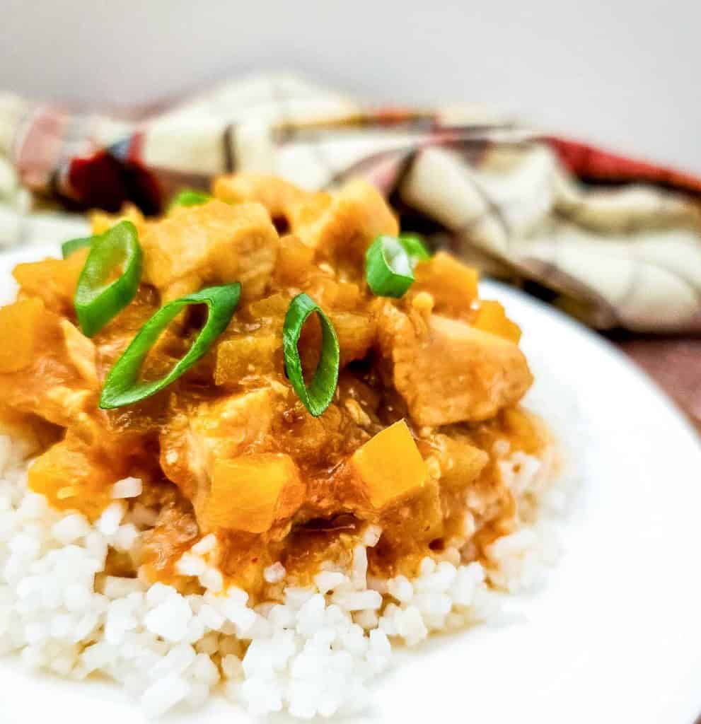 A plate of white rice topped with Crock Pot Hawaiian Chicken in a yellow curry, featuring chunks of meat, diced pineapple, and sliced green onions. A plaid cloth is in the background.