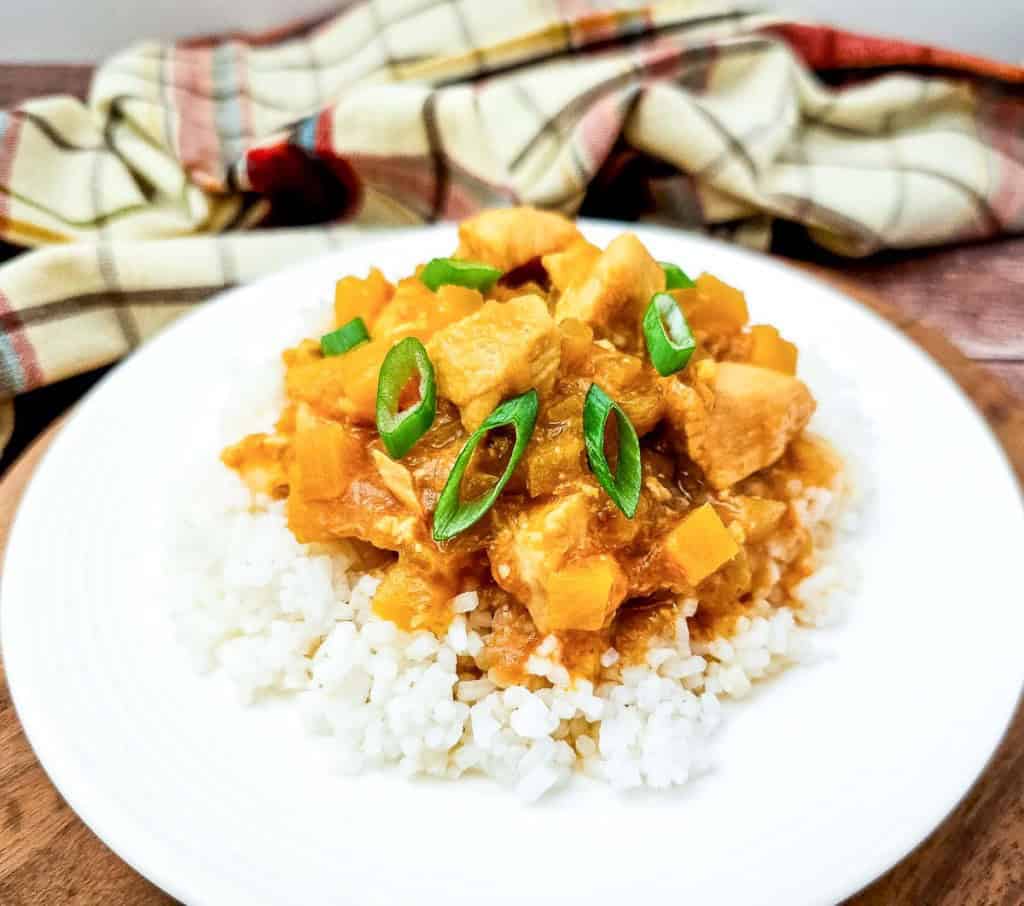 A plate of white rice topped with a savory Crock Pot Hawaiian Chicken stew, featuring diced chicken, yellow vegetables, and green onion slices, with a plaid cloth in the background.