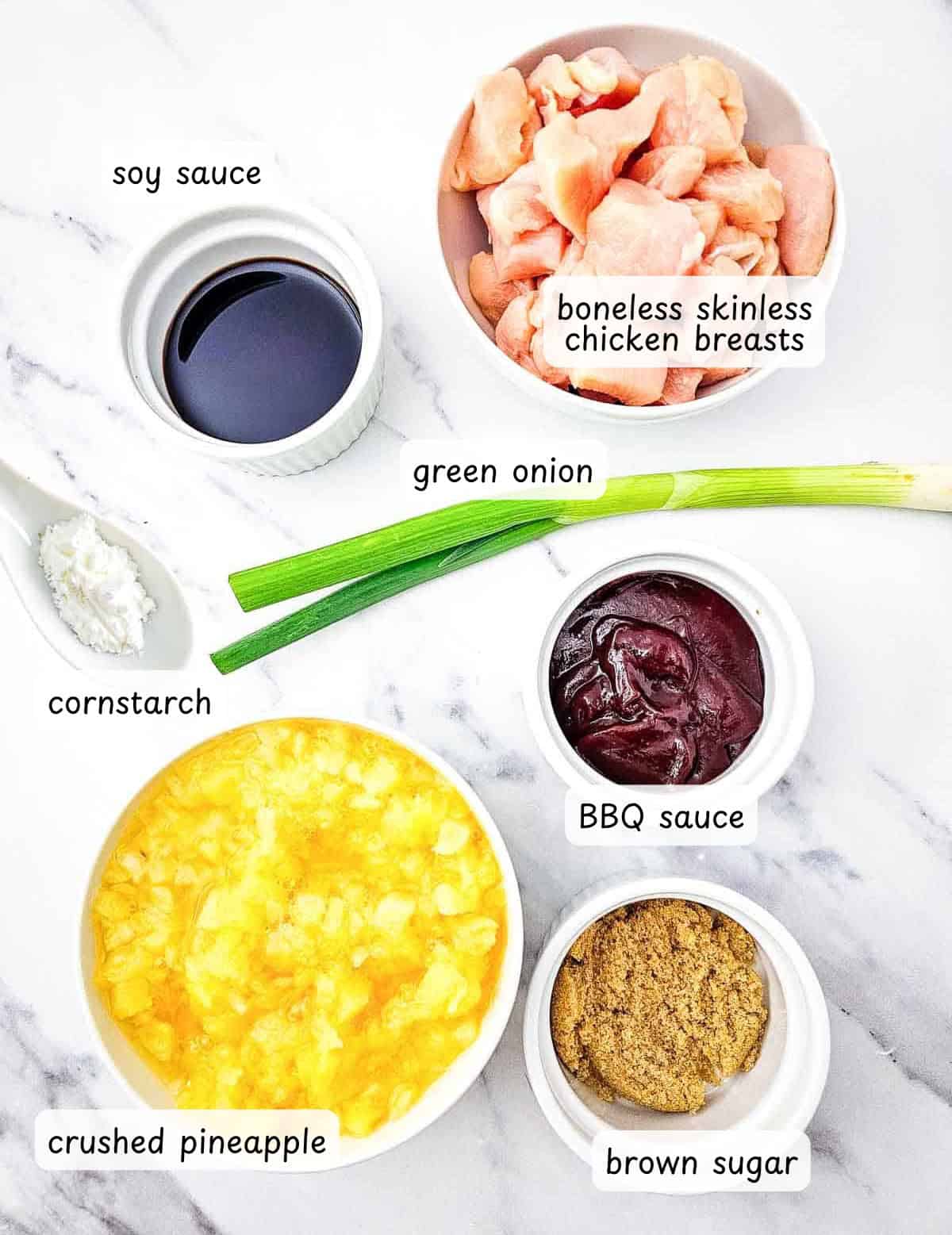 Ingredients for Crock Pot Hawaiian Chicken arranged on a marble surface: soy sauce, boneless skinless chicken breasts, green onion, cornstarch, BBQ sauce, crushed pineapple, and brown sugar.