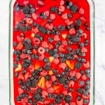 A glass dish brimming with red gelatin is crowned with a medley of berries&mdash;raspberries, blackberries, and blueberries&mdash;evoking the sweet charm of a Mixed Berry Pretzel Salad, all elegantly displayed on a cool marble surface.